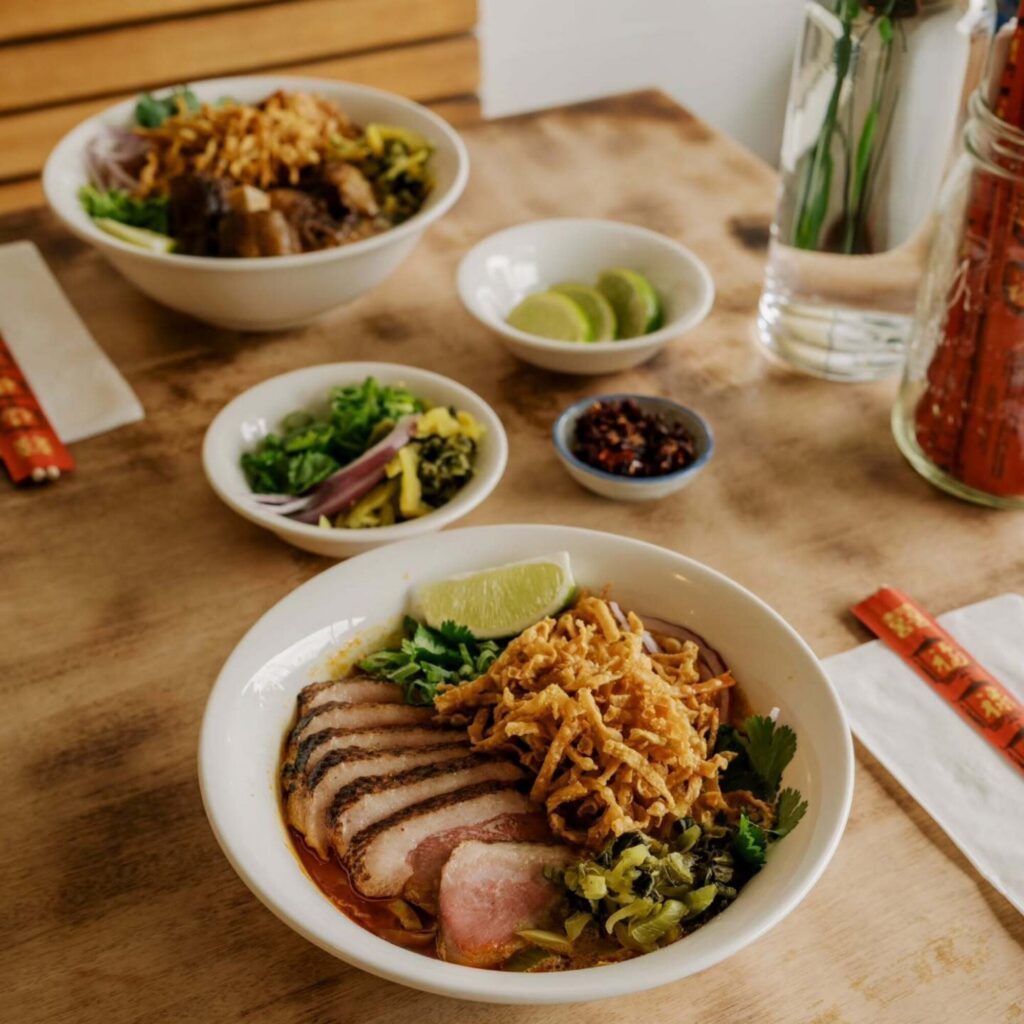 Two bowls of noodle soup with sliced meat and garnishes sit on a wooden table, accompanied by small dishes of lime, pickles, and chili sauce—perfect for seasonal eating and enjoying cozy autumn recipes. Chopsticks and napkins are included.