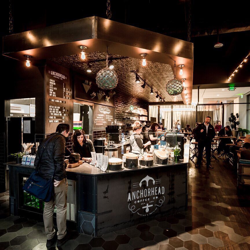 Customers order and baristas prepare drinks at the Anchorhead Coffee bar inside a modern café with warm lighting and hexagonal floor tiles, making it a charming spot for Seattle gifts or a stop on your Mother’s Day Gift Guide.