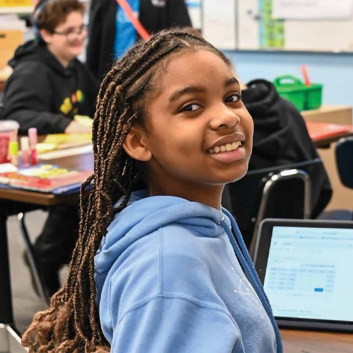 A student wearing a blue hoodie sits at a desk in a classroom, smiling at the camera with a laptop and school supplies, surrounded by classmates—showing there are always more options for learning and connecting.