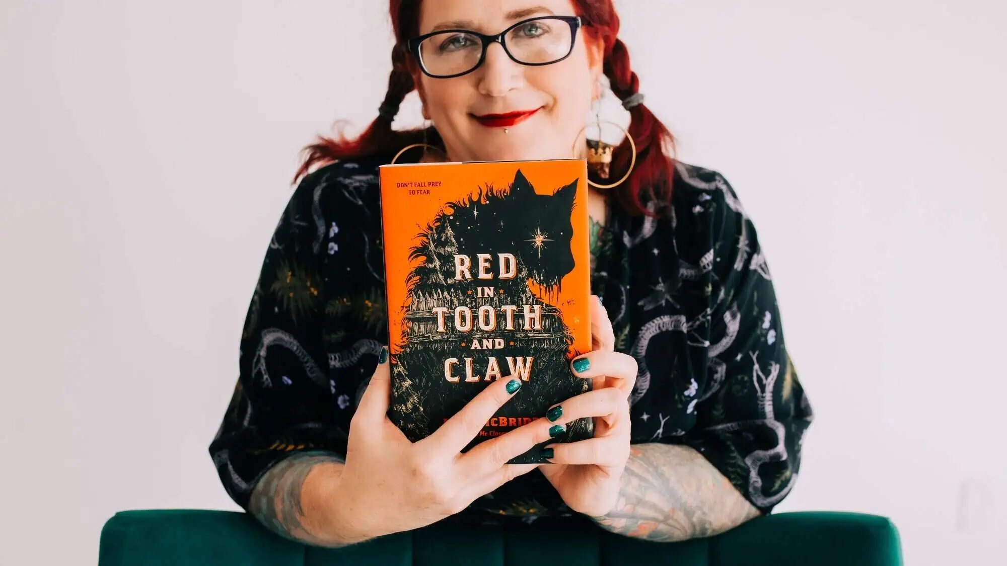 A person with glasses and red hair holds up "Red in Tooth and Claw" by author Lish McBride, a fiction book featuring a black animal silhouette on its orange cover.