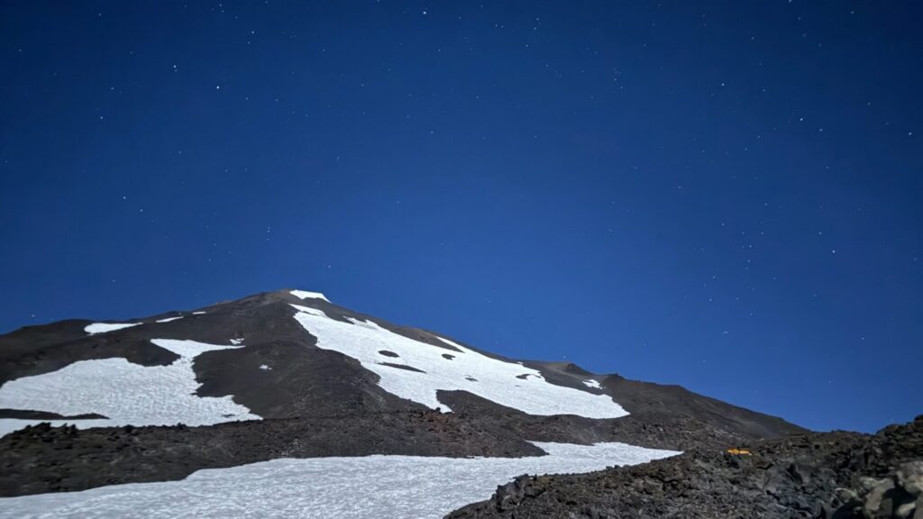 Snow patches cover a rocky mountainside under a clear night sky filled with stars.