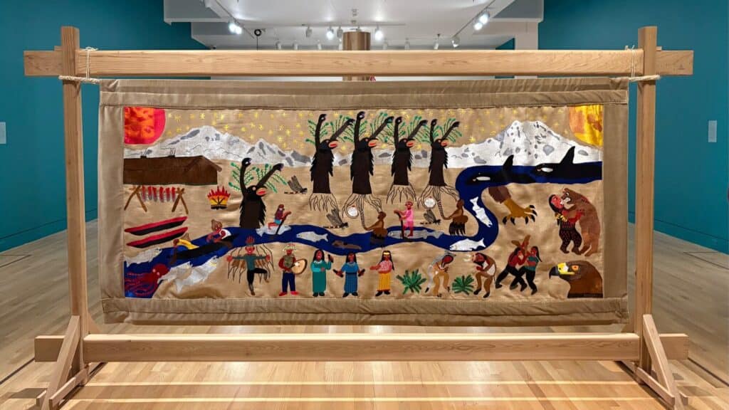 A colorful embroidered textile depicts people, animals, trees, a river, and mountains, displayed on a wooden frame in an art gallery with teal walls and wood flooring.