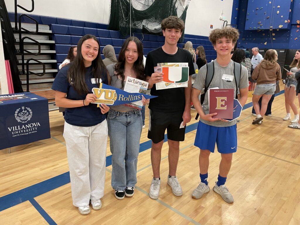 Four UPrep private school students stand in a gym, proudly holding college admission signs for Villanova, Rollins, University of Miami, and Elon. Other people and tables can be seen in the background.