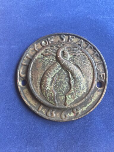 Antique bronze medallion with the city of Seattle seal embossed, featuring a phoenix design, dated 1869, symbolizing the honesty in relationships, placed on a blue background.