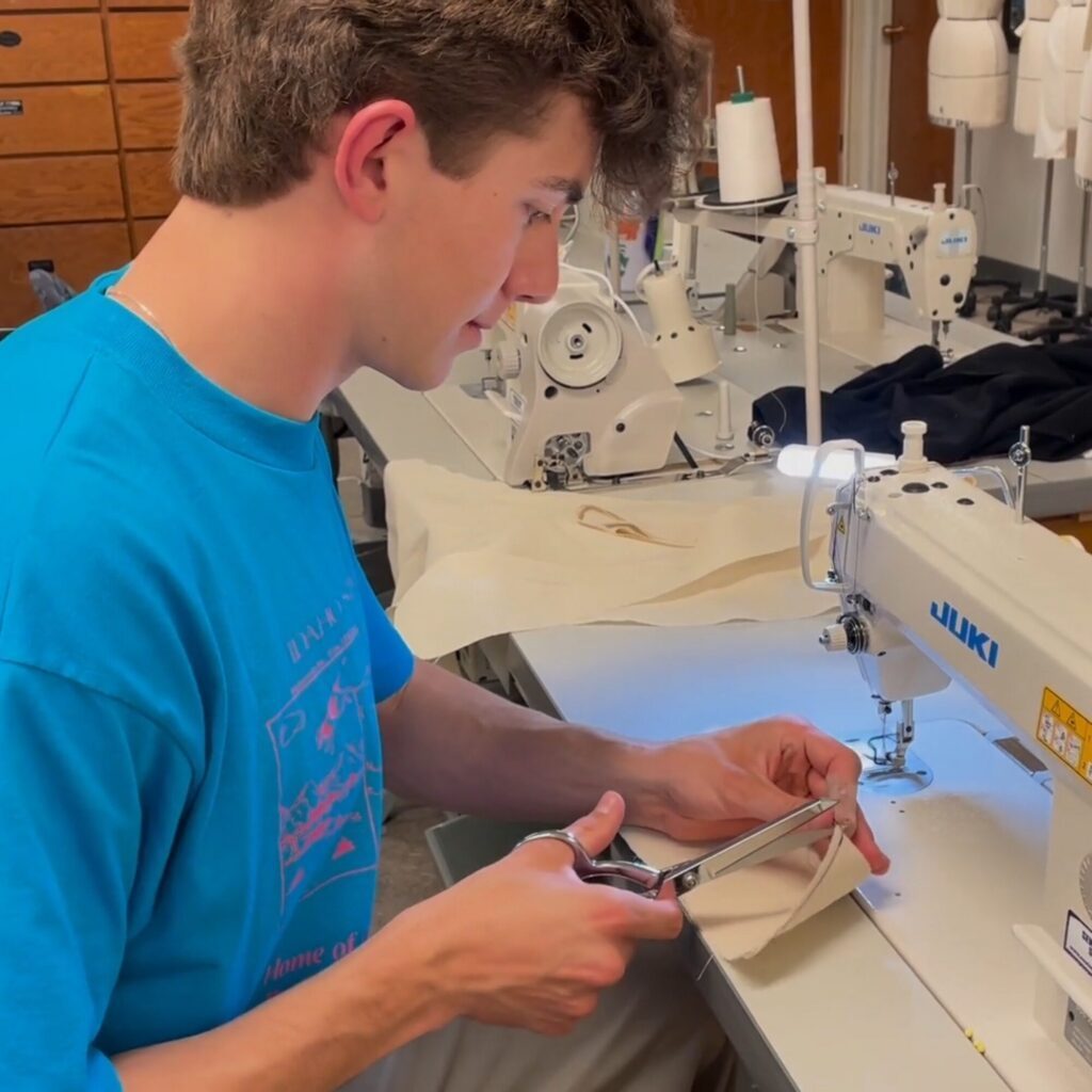 A young fashion designer in a blue shirt uses scissors to cut fabric while sitting at a sewing machine in a workshop, preparing material that may one day appear on the runway.