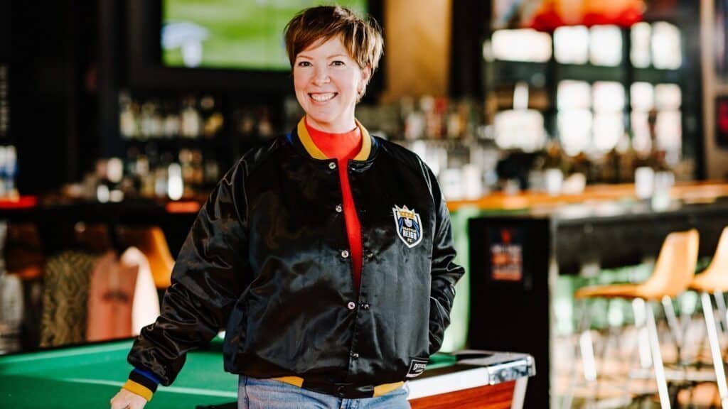 A person stands and smiles beside a pool table in a bar, wearing a black jacket, red top, and jeans. The background shows bar stools and a TV screen.