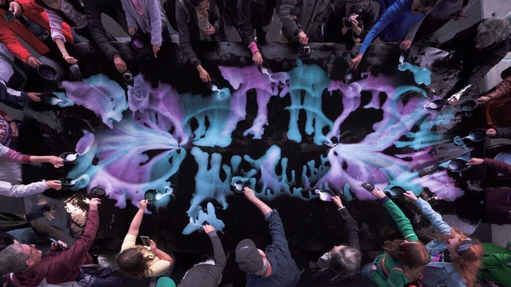 During an art walk in Seattle, people stand around a large black table, creating symmetrical patterns with blue and purple powders—colors that celebrate the vibrant LGBTQIA+ community—as they spread them outward with spoons.