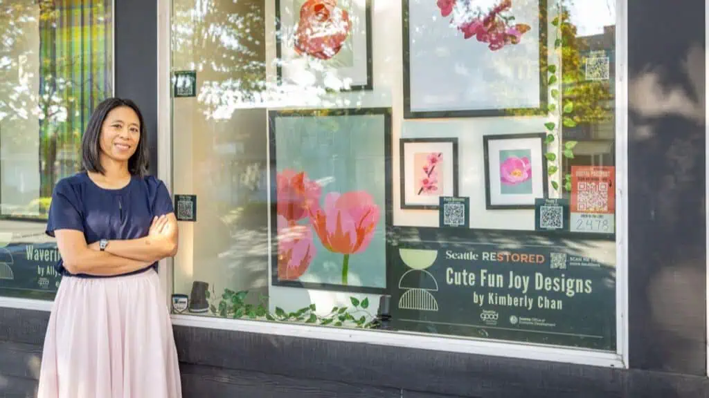 A woman stands smiling with arms crossed beside a shop window displaying floral artwork and a sign for "Cute Fun Joy Designs by Kimberly Chan.