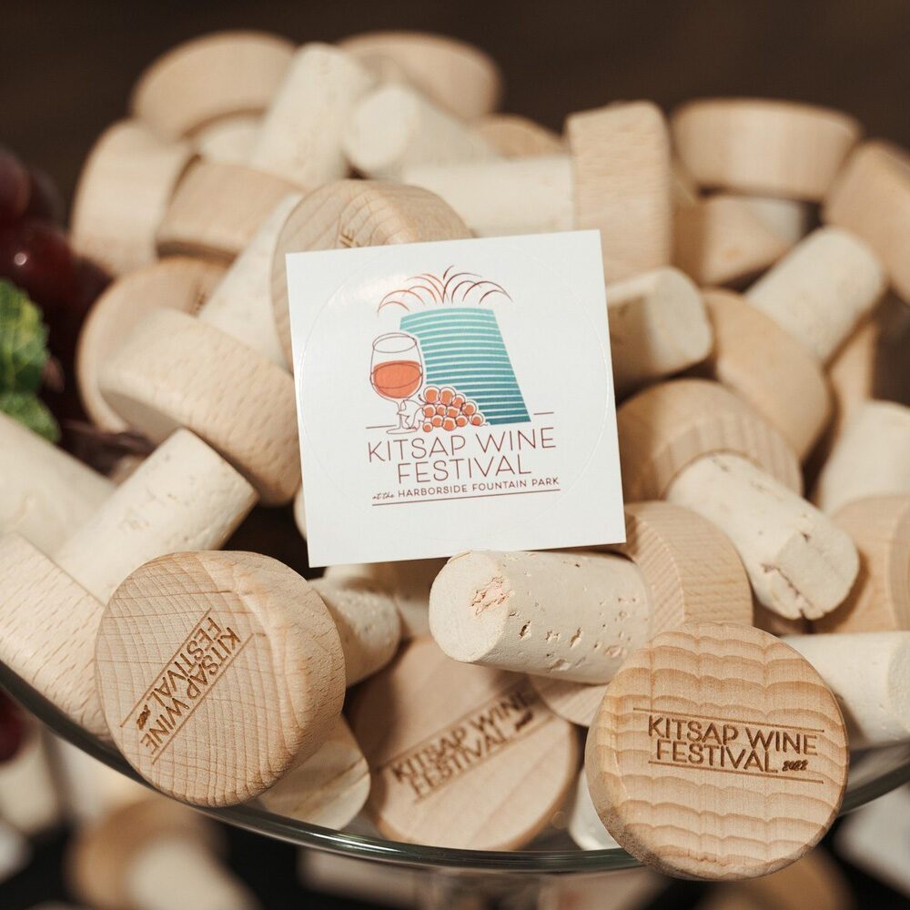 A bowl filled with wine corks, some stamped with "Kitsap Wine Festival," and a sticker displaying the festival's logo, event details, and the Must List date: 08-07-2025.