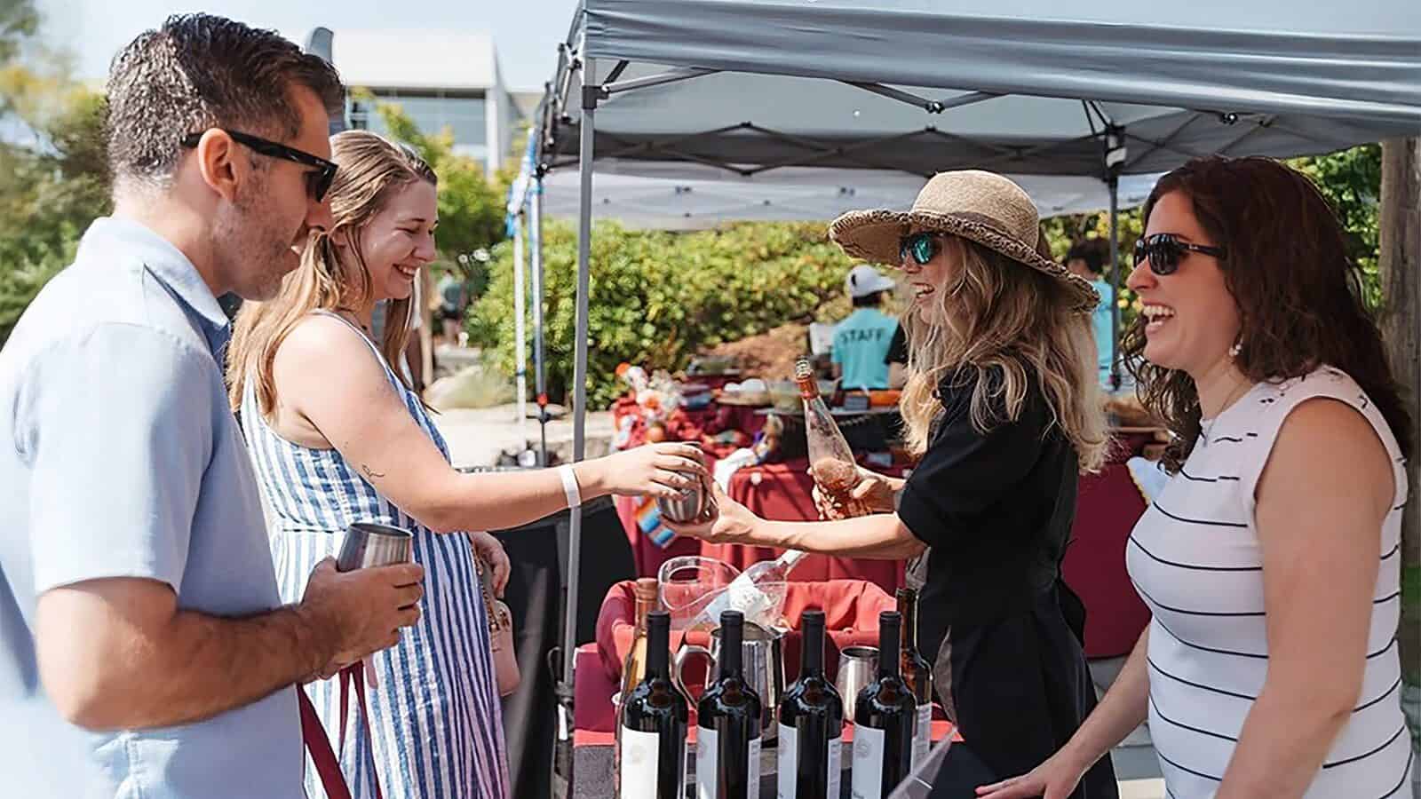 People at the South Lake District Block Party stand by a table with bottles of wine, smiling and interacting under a canopy on a sunny day—a perfect addition to your list of what to do in Seattle this week.