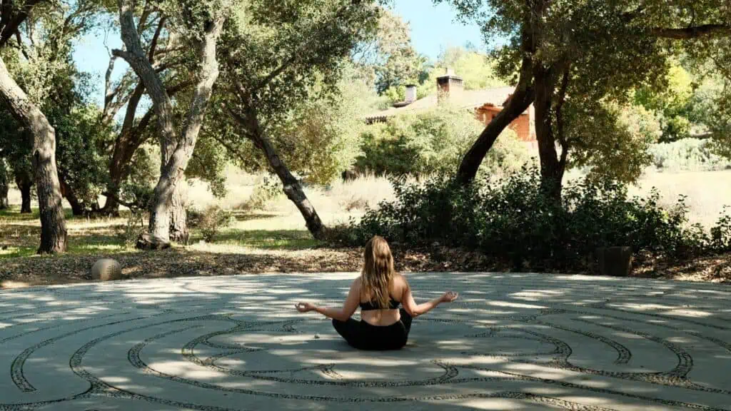 A woman sits cross-legged in meditation on a stone labyrinth outdoors at Rancho La Puerta, surrounded by trees and a house, embracing the serenity of this holistic health sanctuary.
