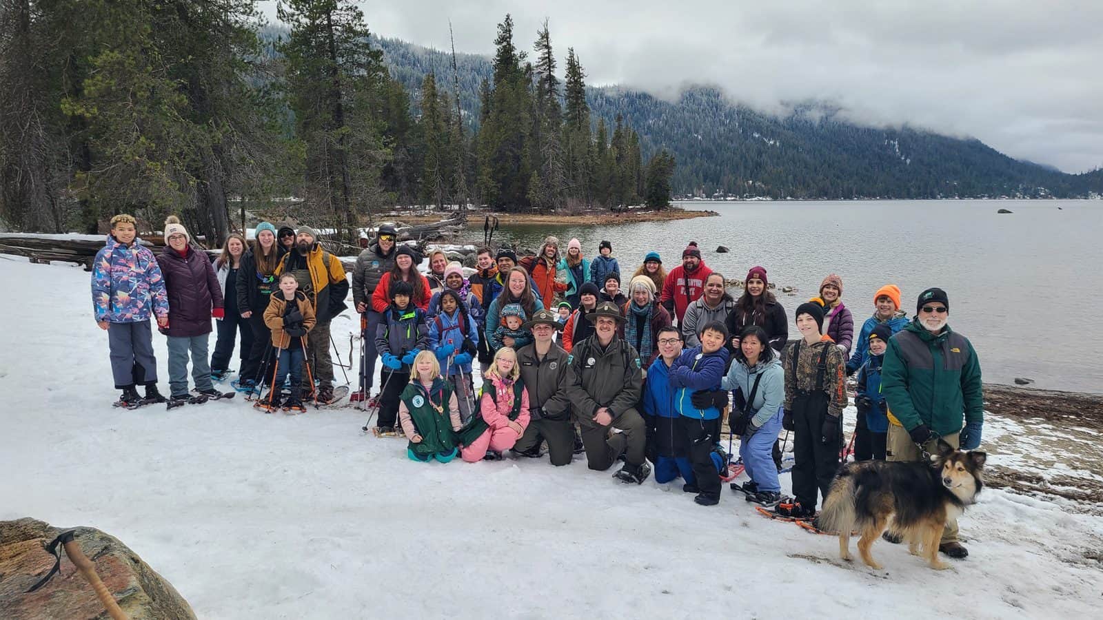 A large group of people and a dog stand on a snowy lakeshore, embracing "A Seattle New Year For All Types," surrounded by trees and majestic mountains.