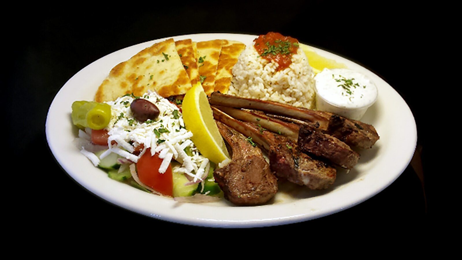 A plate of comfort food featuring grilled lamb chops, rice, pita bread, Greek salad with feta, a lemon wedge, and a side of white sauce, served on a white oval platter against a black background.