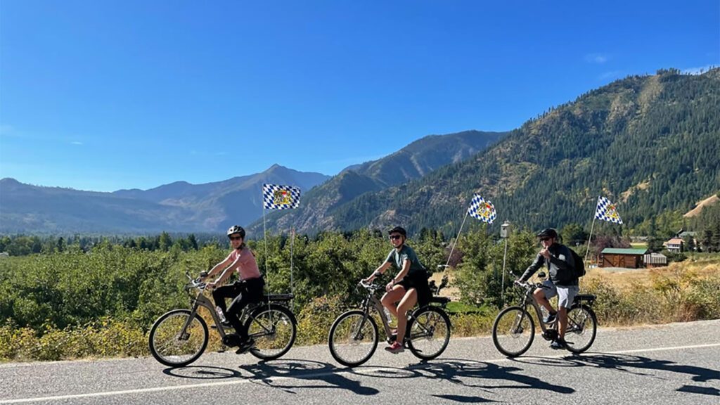 Three people ride bicycles on a road with scenic mountains and greenery in the background, under the clear blue sky of Hidden Washington's Leavenworth, perfect for exploring all year.