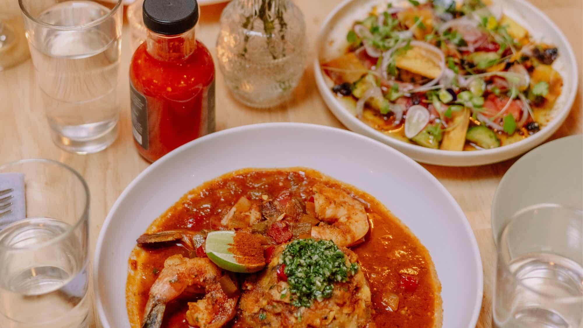 A plate of seafood in tomato sauce with herbs, a side plate of salad with sliced vegetables, and bottles of sauce on a wooden table—perfect for fall recipes and cozy seasonal eats.