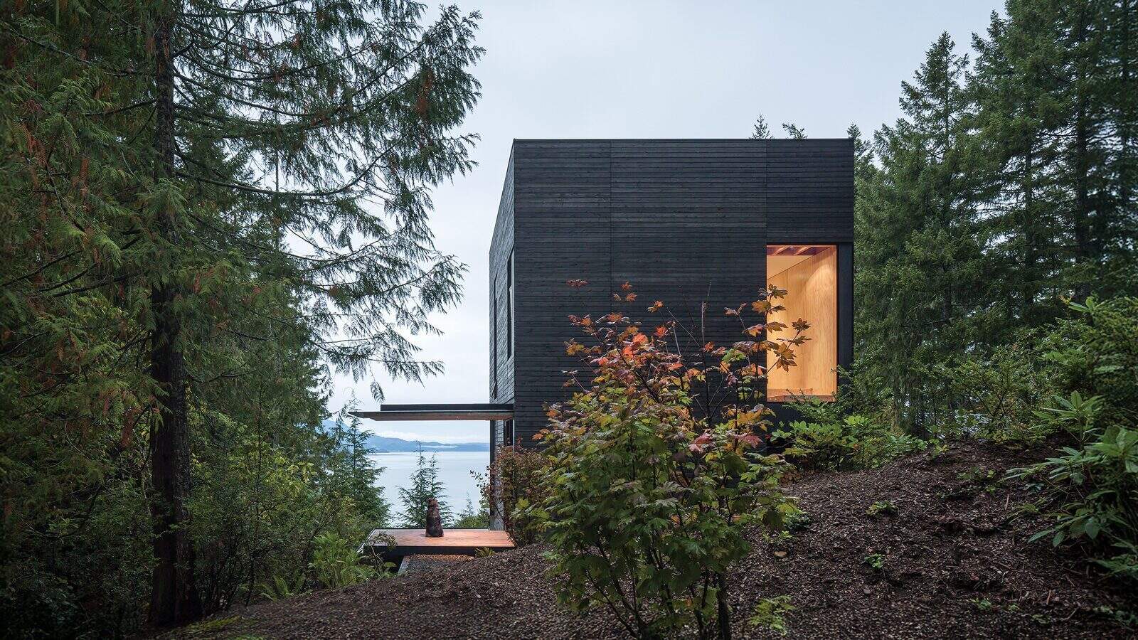 A modern, dark wood cabin with large windows sits on a forested hillside—ideal for second homes or summers in the waterfront—partially obscured by trees and foliage, overlooking the water in the background.