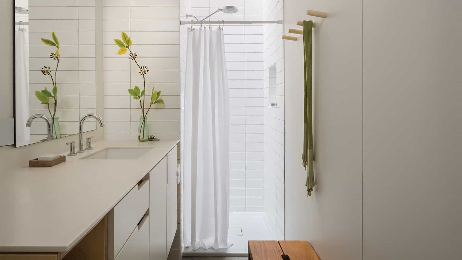 A modern bathroom with a white countertop, dual sinks, and a shower with a white curtain evokes relaxing summers in the waterfront. Green towels and two glass vases with leafy branches add freshness—perfect for second homes.