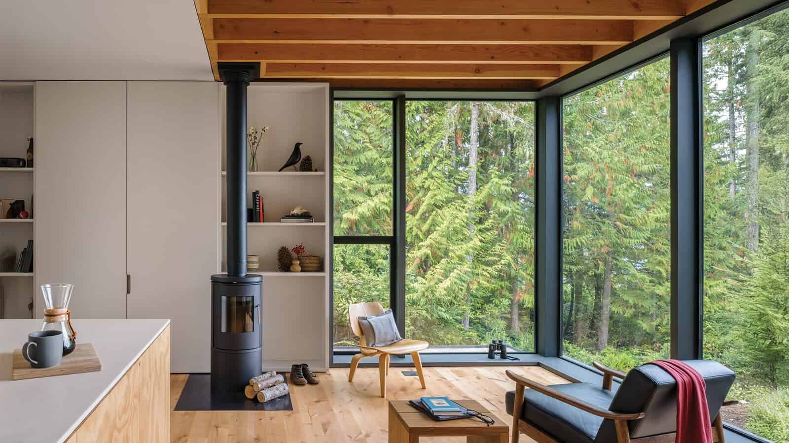 A modern living room with a wood stove, wooden chair, coffee table, and large floor-to-ceiling windows overlooking a forest—perfect for second homes or enjoying relaxing summers in the waterfront.