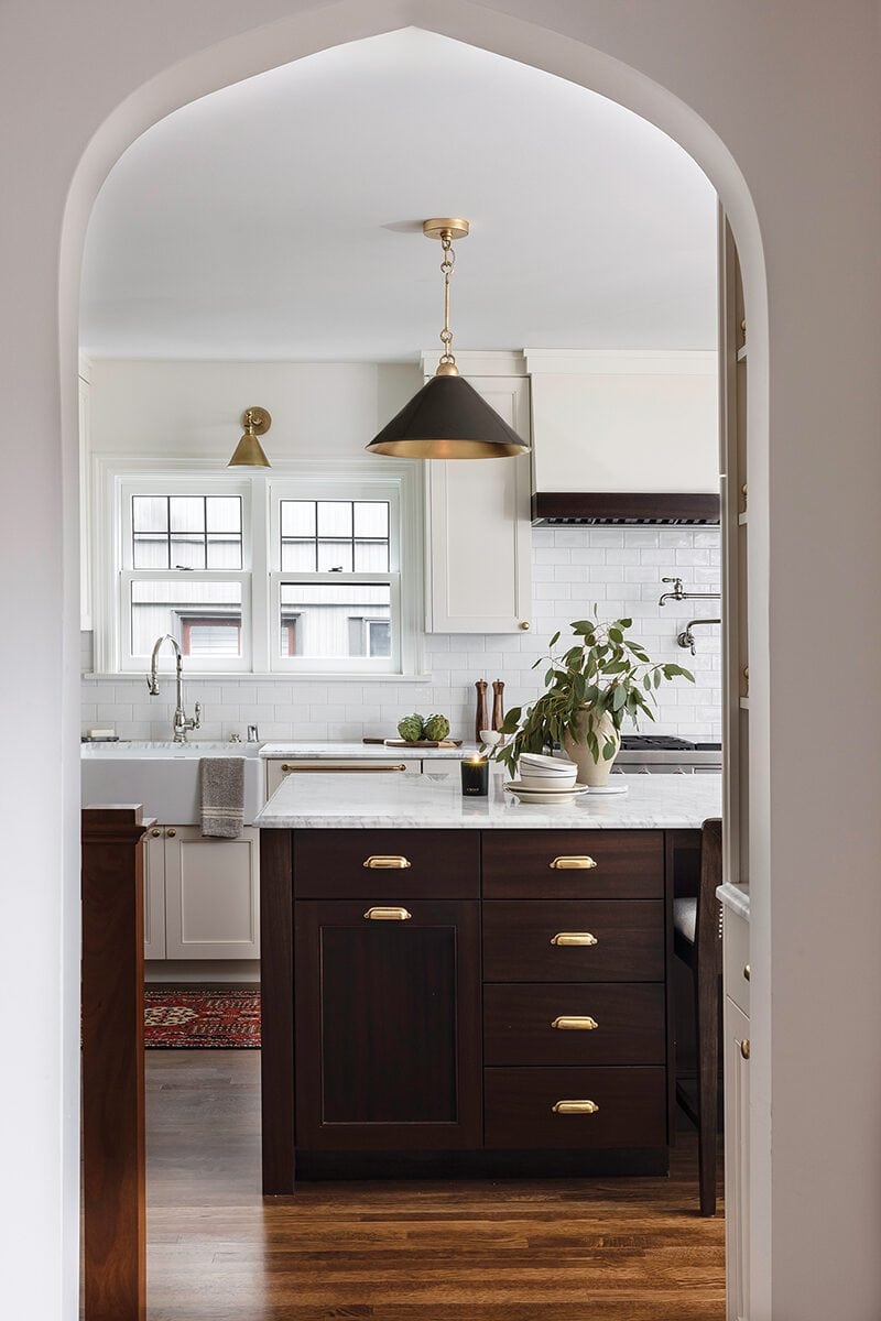 View through a Tudor-style arched doorway into a kitchen with white cabinets, a dark wood island, brass fixtures, and a potted plant on the counter. Natural light pours through two windows over the sink, capturing the charm of living in Montlake.