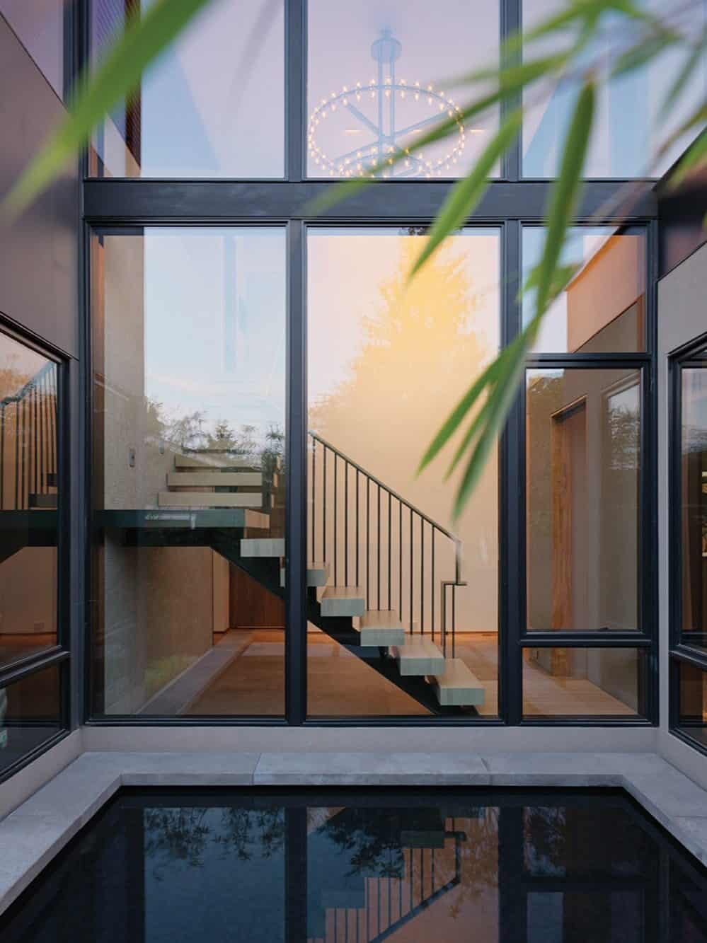 Modern glass-walled interior with a floating staircase, black metal railings, a pendant chandelier, and a reflection pool in the foreground—impeccable details that reflect the sophistication of custom-built homes.