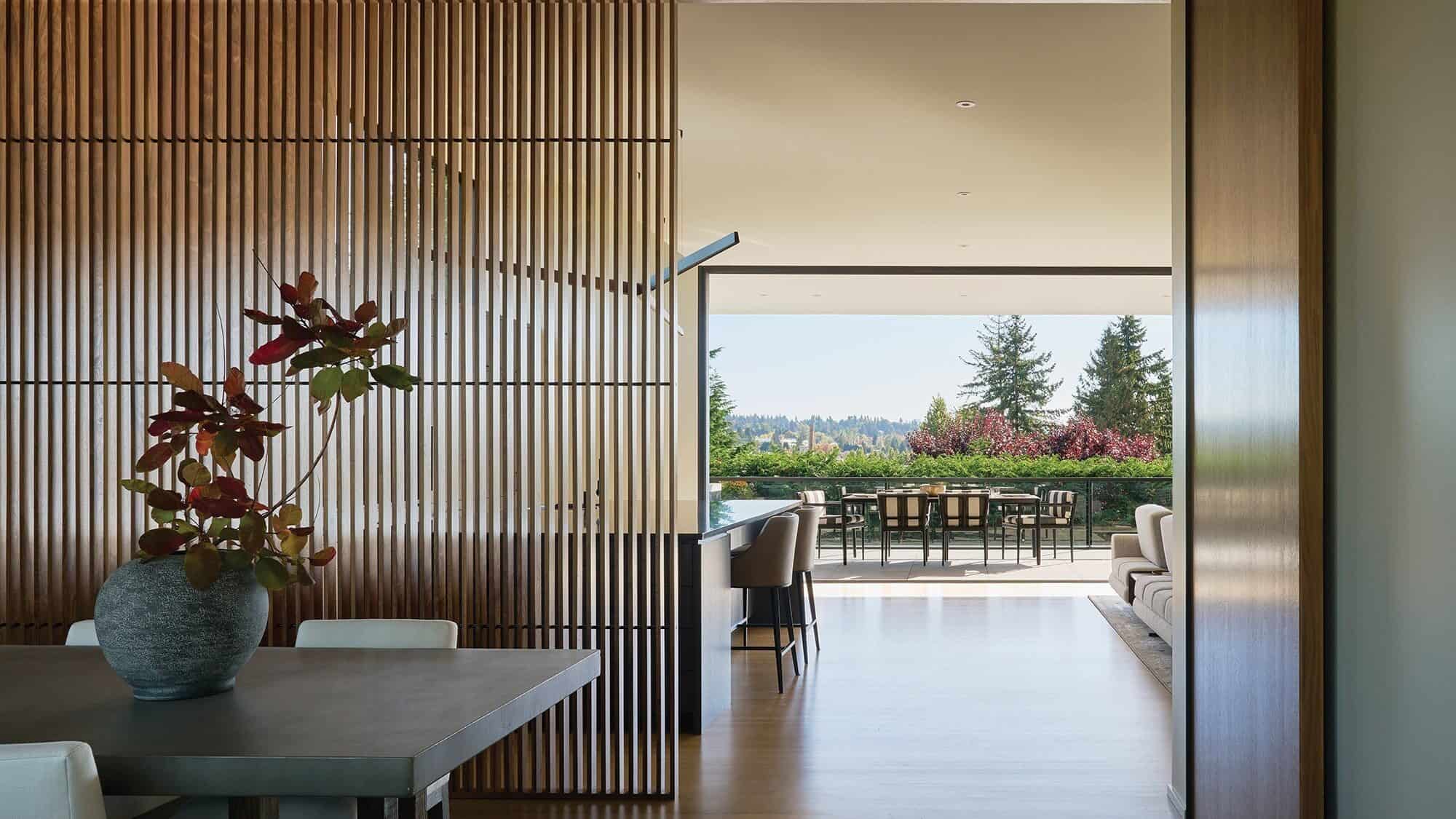 Modern open-plan living and dining area with wooden slat partition, vase with red leaves, and large windows overlooking trees and an outdoor patio—perfect for those seeking the comfort and style of custom-built homes.
