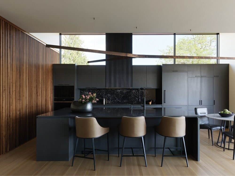 Modern kitchen with dark cabinetry, black marble backsplash, island with three tan barstools, wood panel accent wall, and large windows letting in natural light—perfect inspiration for custom-built homes.