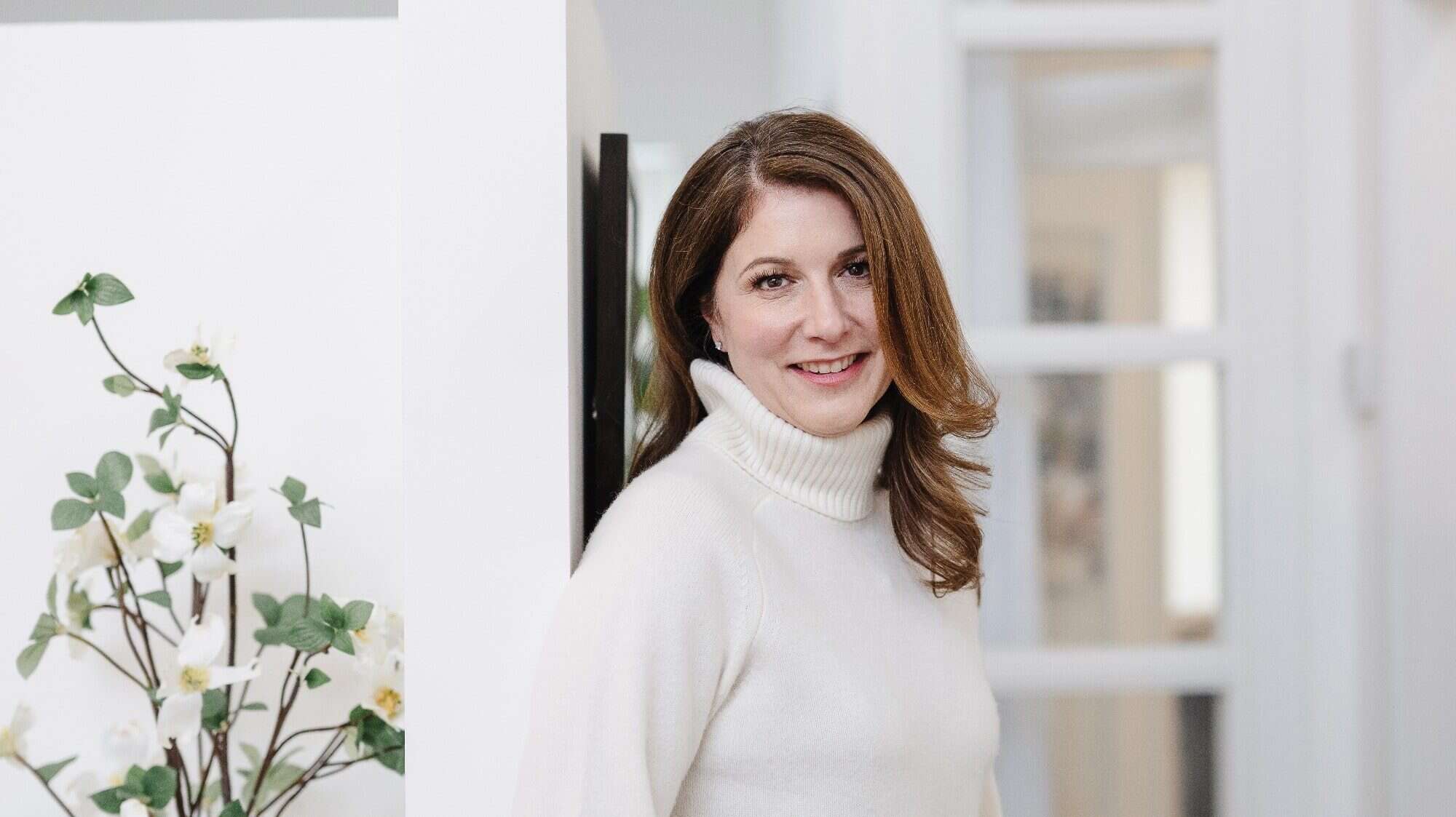 A woman in a white turtleneck sweater leans against a wall near a doorframe, embodying the Seattle home design aesthetic. A plant with white flowers graces the foreground, adding a touch of nature to this elegantly curated interior design space.