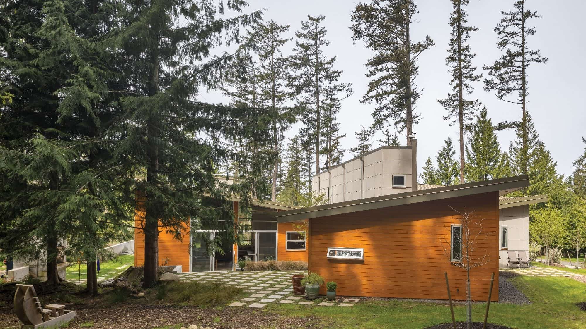 Modern house with wood siding and large windows surrounded by tall trees, embodying the Seattle home design aesthetic. A stone pathway and small garden are in the foreground, adding charm to the exterior that complements an interior design focused on natural light.