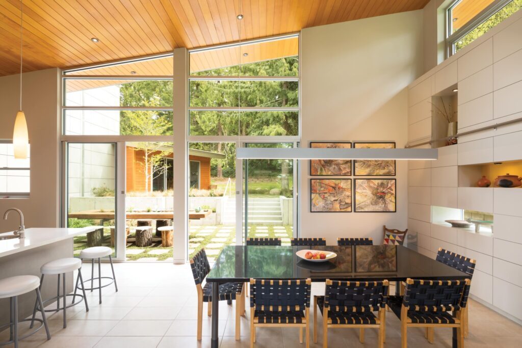 Modern dining area showcasing the Seattle home design aesthetic with a large glass window, black table, six chairs, and wall art. Natural light highlights the wooden ceiling and white walls, complementing the interior design. Enjoy a serene garden view outside.