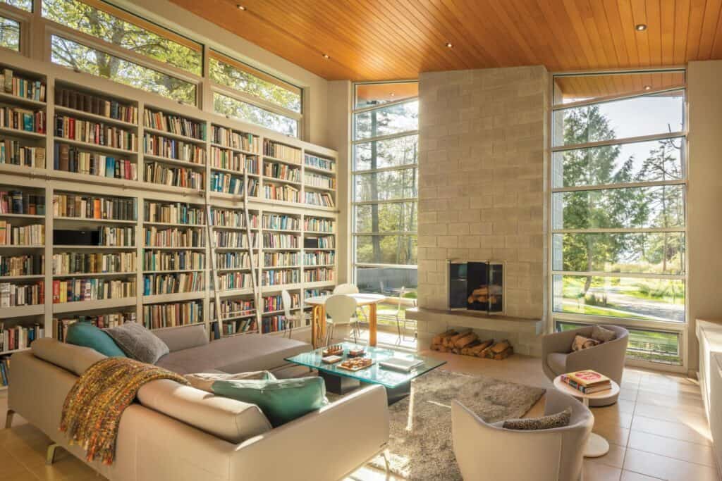 A bright, modern living room reflecting that distinct Seattle home design aesthetic features a large bookshelf, a fireplace, and comfortable seating. Floor-to-ceiling windows offer a view of trees outside.