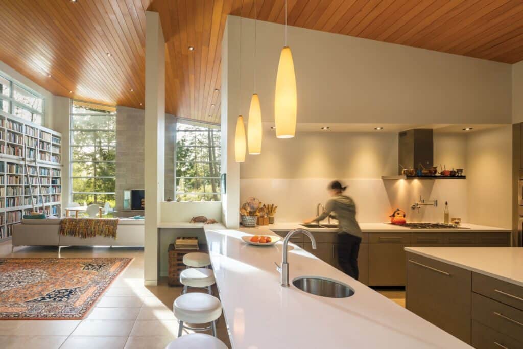 Modern kitchen with a large island, pendant lights, and a person at the sink. The space showcases Seattle home design aesthetic, blending seamlessly with an adjacent cozy living area featuring bookshelves and expansive windows.