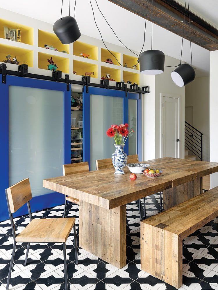 This dining area in a new home built in the 1990s features a wooden table with benches, black and white geometric floor tiles, blue sliding doors, sleek hanging black lights, and a vase of red flowers as the centerpiece.