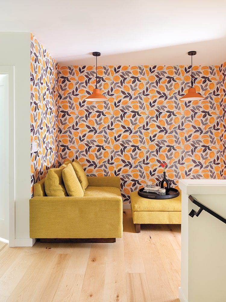 This cozy room in a new home built in the 1990s features orange and yellow floral wallpaper, a cheerful yellow sofa with a matching ottoman, light wood flooring, and two vibrant hanging orange lamps.