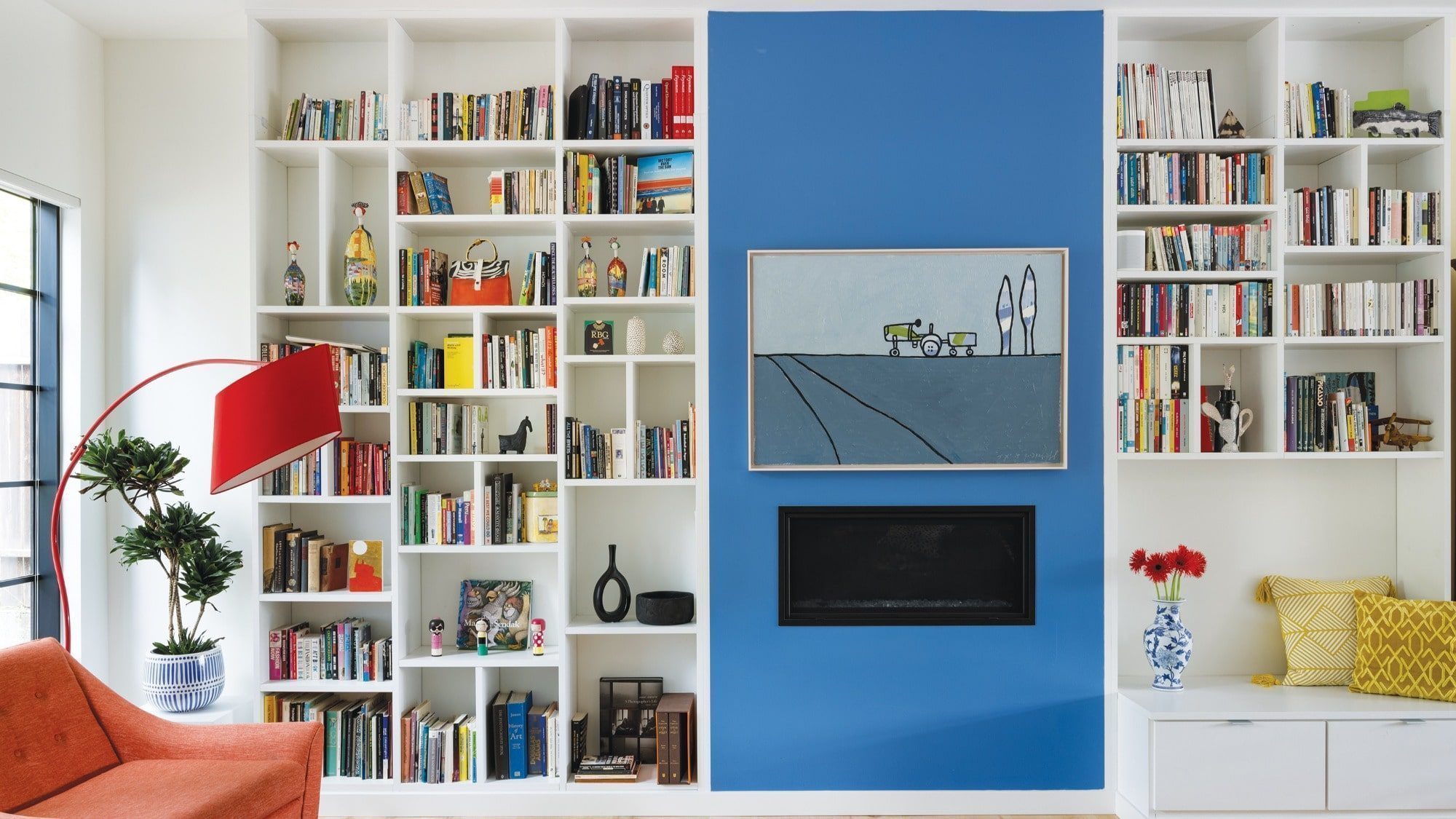 This cozy room, part of a new home built in the 1990s, features white bookshelves that frame a striking painting on the blue wall. A vibrant red floor lamp stands beside an orange chair, with decorative items accenting the space. The fireplace adds warmth and character to this inviting nook.