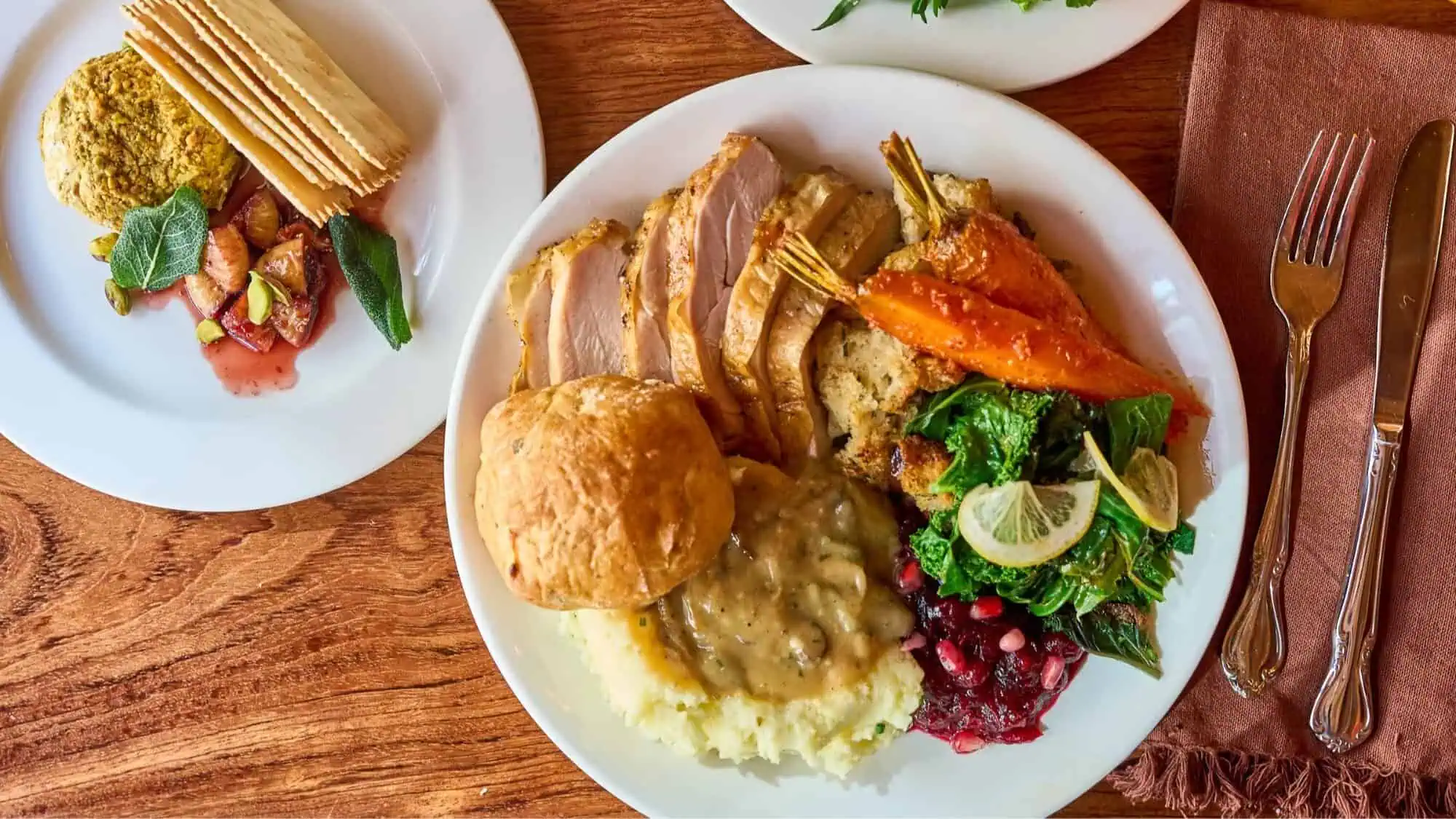 A plate with sliced turkey, stuffing, a bread roll, mashed potatoes with gravy, roasted carrots, sautéed greens with lemon, and pomegranate seeds, next to a plate of crackers and cheese.