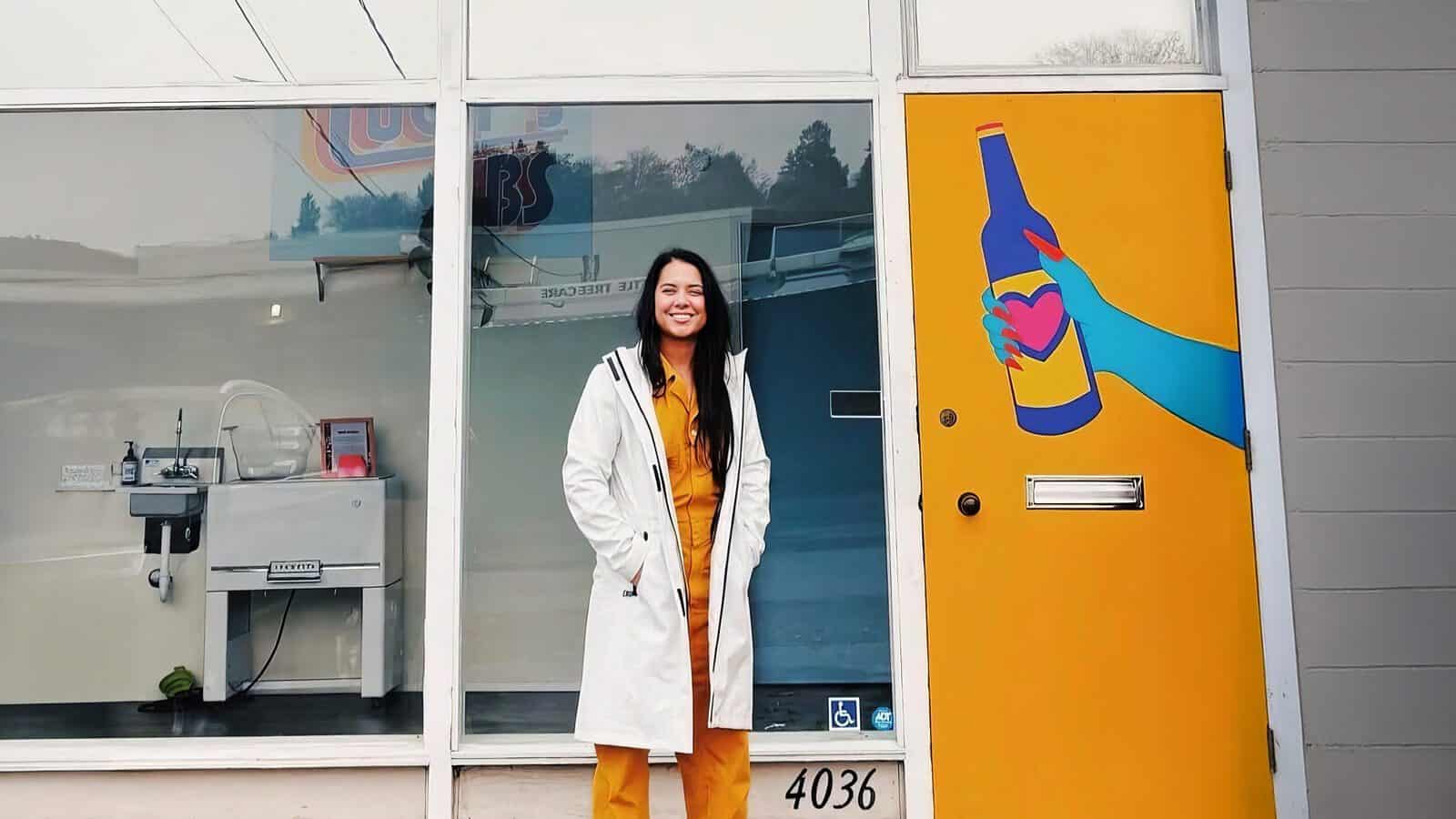 A woman in a white coat and yellow outfit stands smiling in front of a storefront with a yellow door painted with a hand holding a bottle, celebrating Lucy’s Lifts Up Underrepresented Winemakers.