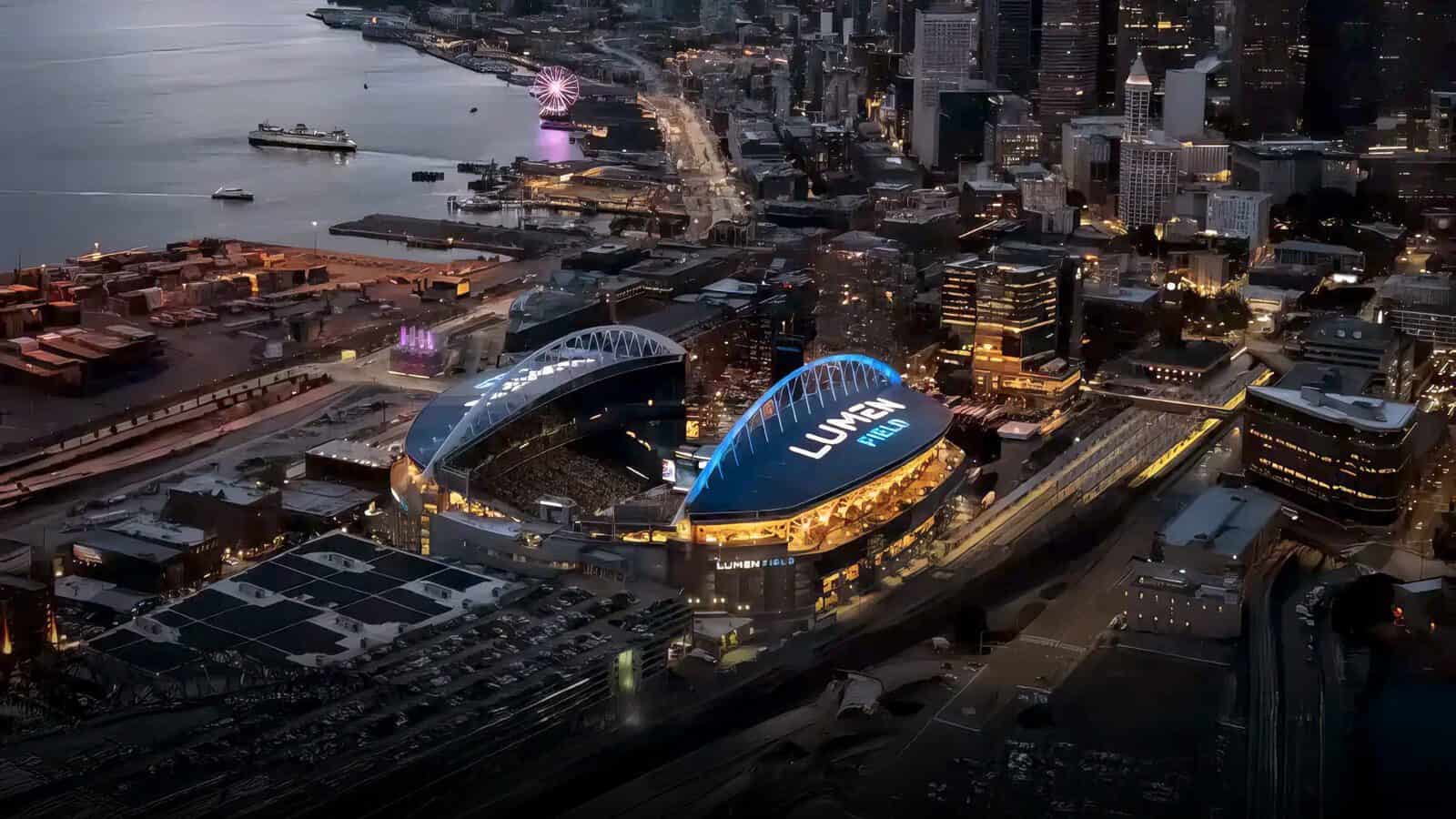 Aerial view of Lumen Field stadium illuminated at night in downtown Seattle, with adjacent waterfront, city buildings, and nearby streets visible.