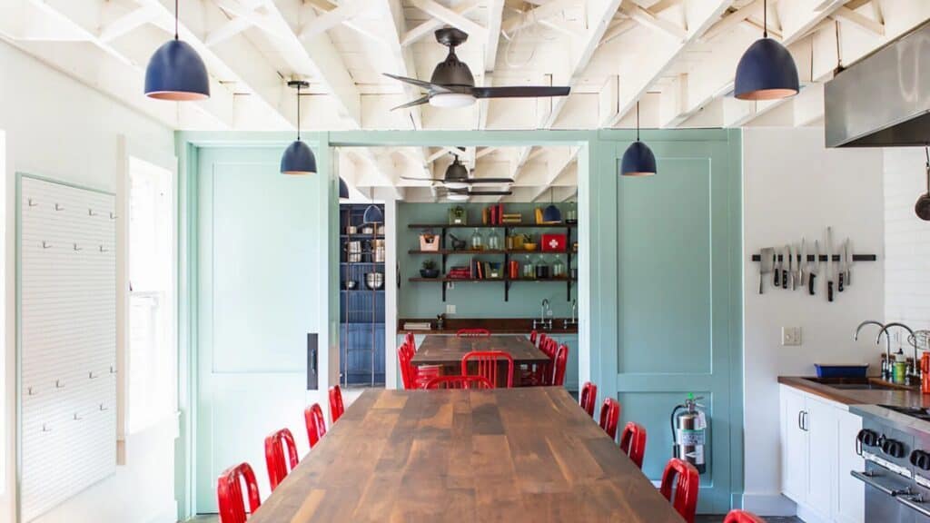 A spacious dining area with a long wooden table, red chairs, exposed white ceiling beams, blue pendant lights, and a visible kitchen area to the right—perfect for discussing art and food during the impressionism era.