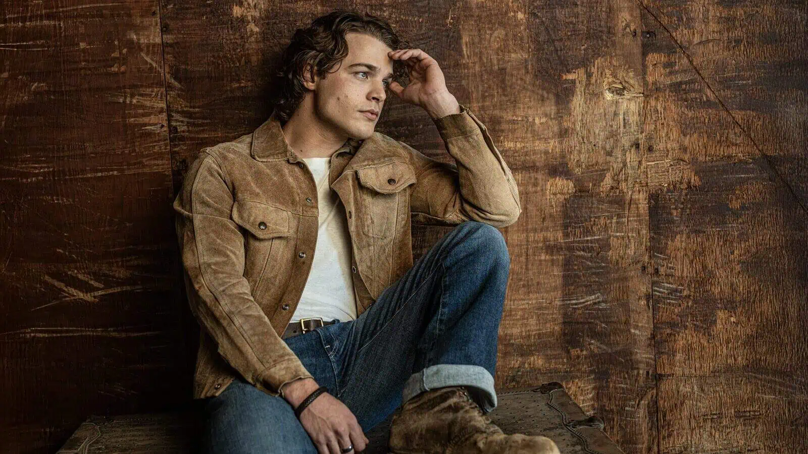 A man in a tan suede jacket, white shirt, and jeans sits on a wooden surface against a rustic wooden wall, looking thoughtfully into the distance.
