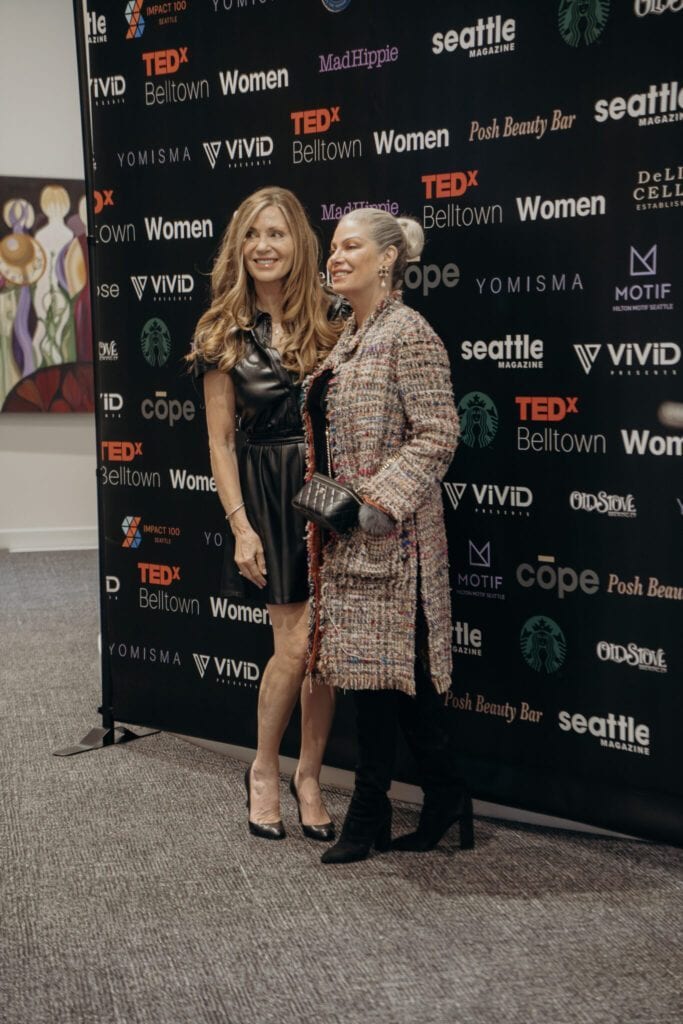 Two women pose and smile together in front of a step-and-repeat banner at an indoor event.