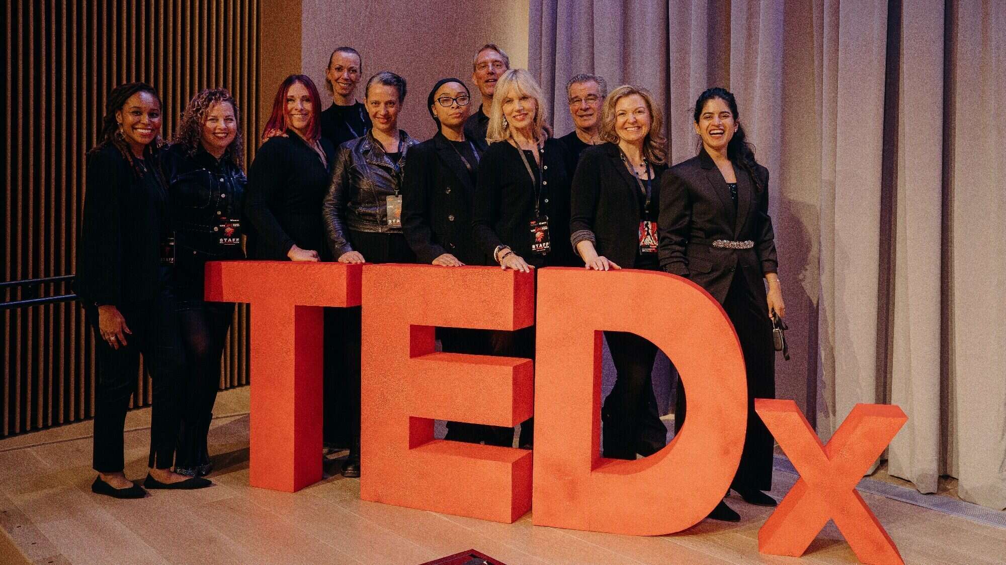 A group of twelve people stands behind a large red TEDx sign on a stage, posing for a group photo.