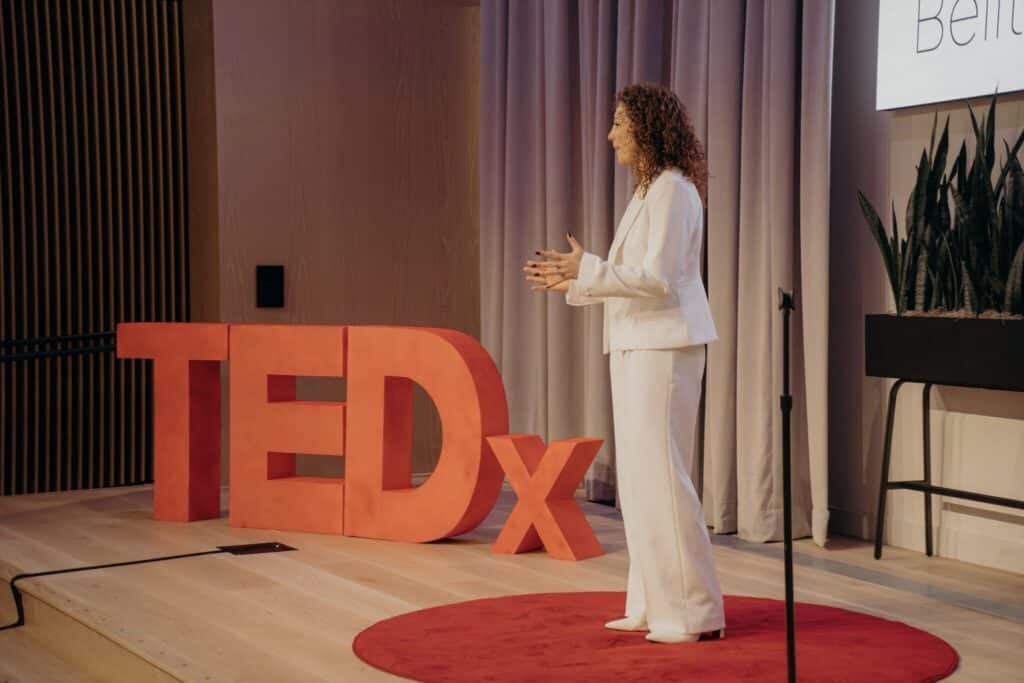 A woman in a white suit stands on a red circular carpet, speaking at a TEDx event with a large red TEDx sign behind her.