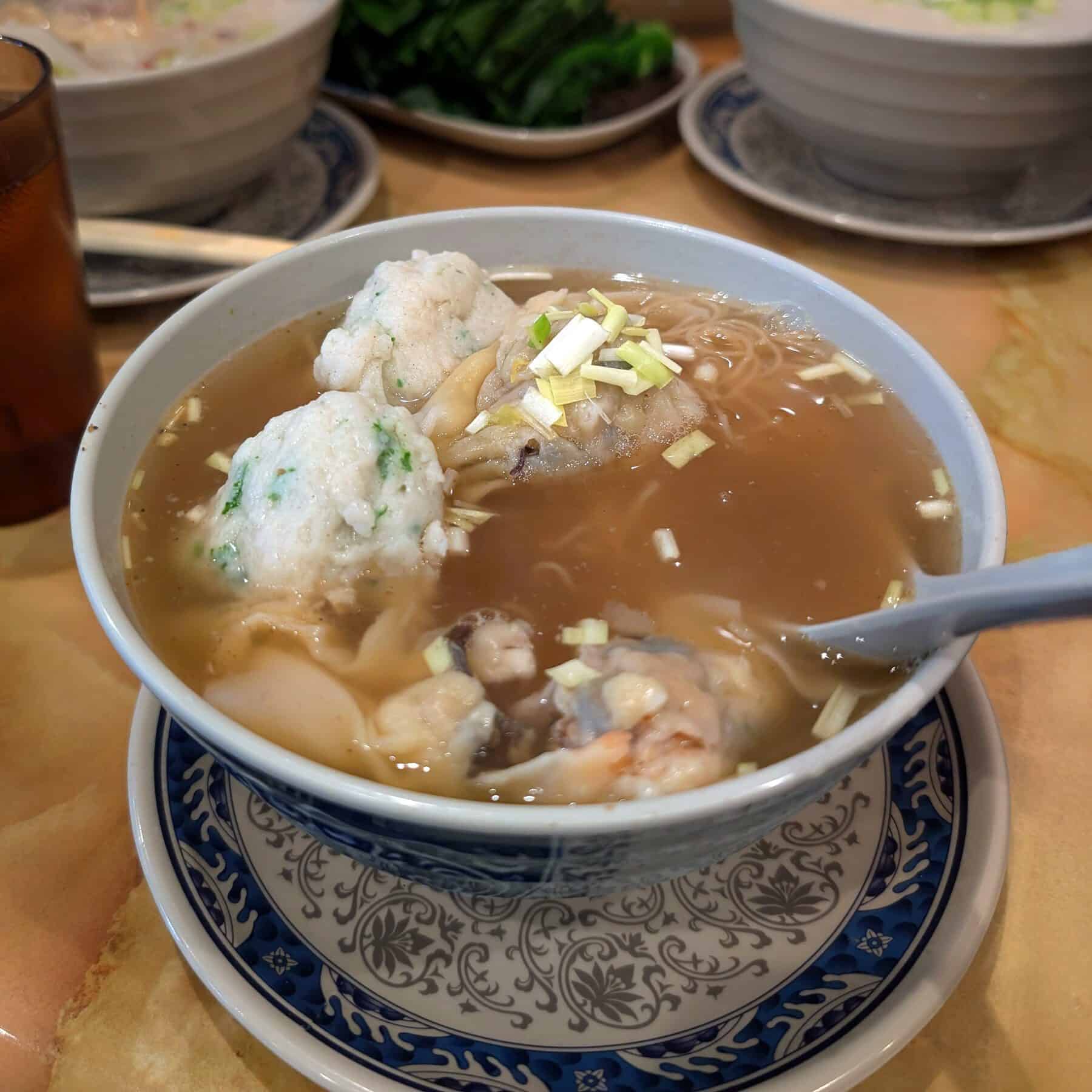 A bowl of noodle soup with fish balls, vegetables, and chopped green onions, perfect for September recipes, served on a decorative plate with a spoon on the side.