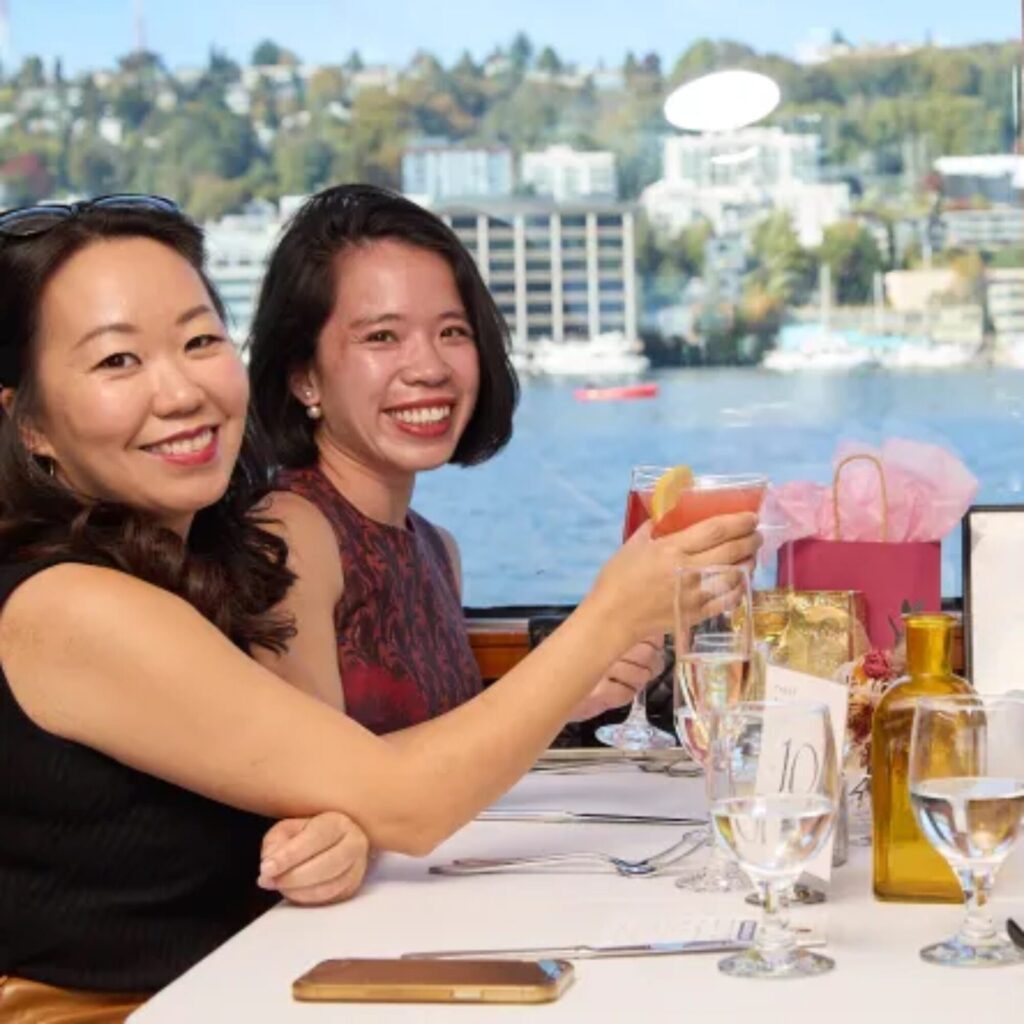 Two women sit at a table by a window, smiling and holding drinks, with a cityscape and water behind them—capturing the perfect moment for a Seattle Mother’s Day gifts inspiration or your next Seattle gift guide.