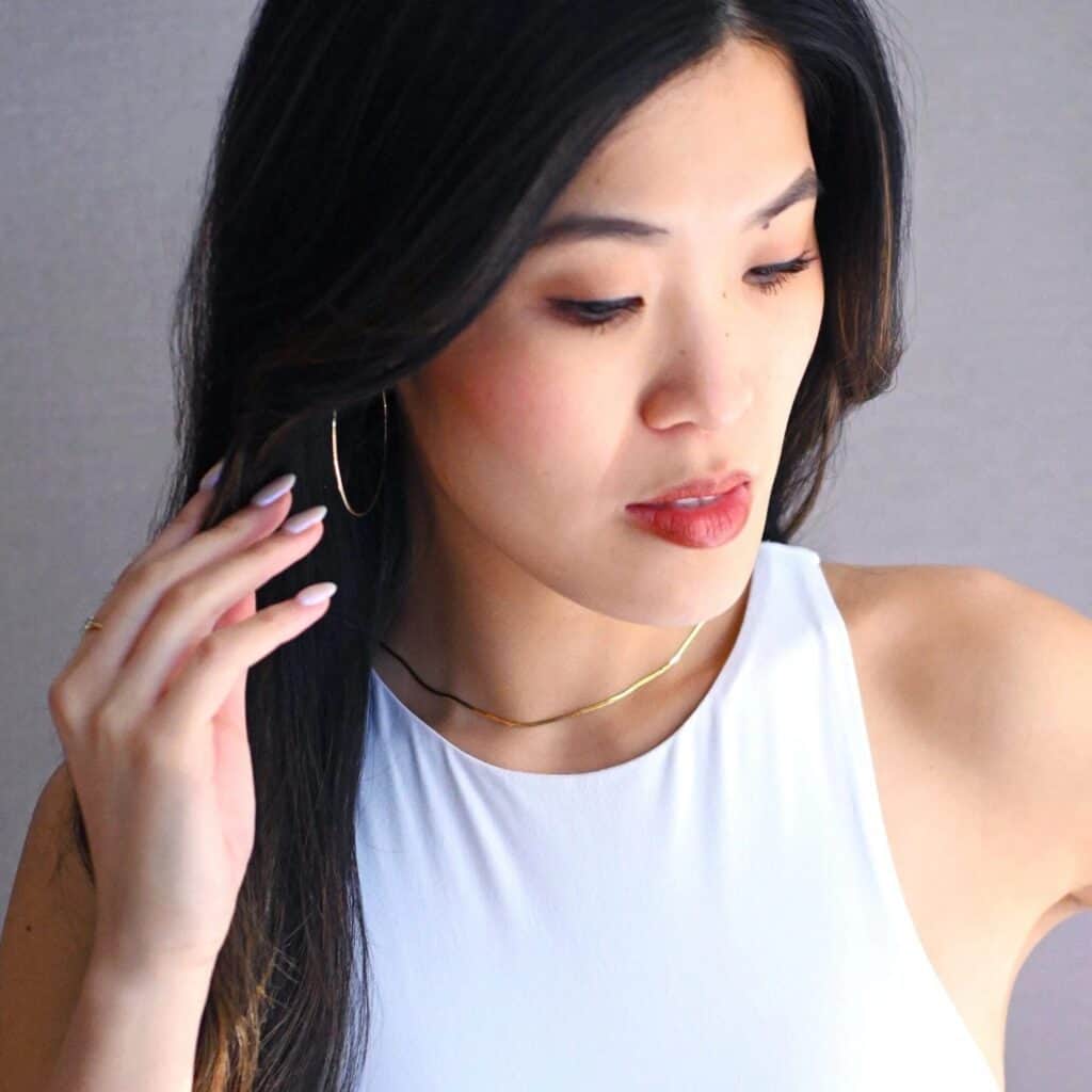Woman with long dark hair wearing a white sleeveless top, gold hoop earring, and a gold necklace, looking down and touching her hair—perfect inspiration for your Seattle Mother’s Day gifts or exploring the ultimate Mother’s Day Seattle style.