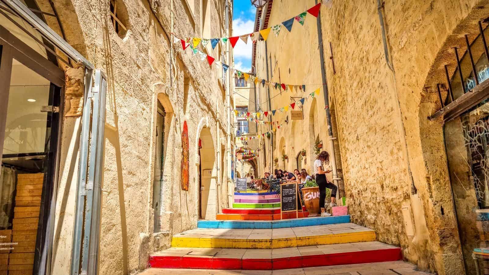 Hidden within one of the Ten Top Travel Destinations from Seattle for 2025, this narrow alley boasts colorful steps, vibrant flags, and cozy outdoor seating nestled against historic stone buildings under a bright blue sky.