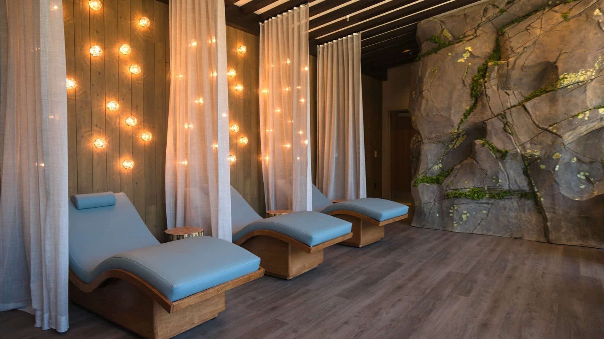Three blue cushioned lounge chairs sit in a spa room with wood floors, sheer curtains, wall lights, and a rock wall with greenery—perfect for unwinding after gaming or dining at the nearby casino.