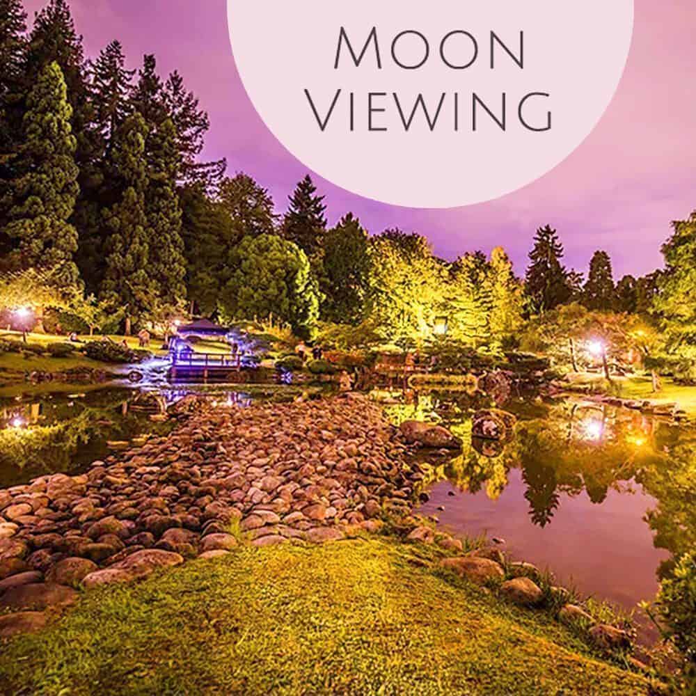 Illuminated Japanese garden at night with a stone pathway over a pond, surrounded by trees beneath a purple sky. "Moon Viewing"—a Must List experience for 09-04-2025.