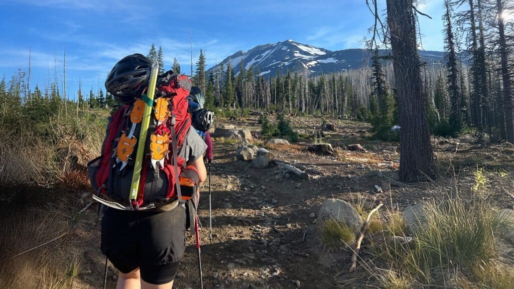 A hiker with a large backpack and trekking poles walks on a rocky trail toward a snow-capped mountain surrounded by trees.