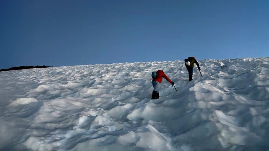 Two people wearing helmets and backpacks use ice axes to climb a steep, snowy, and uneven slope under a clear blue sky.
